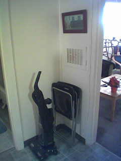 An upright vacuum and two plastic folding chairs in a corner, with a small painting above them